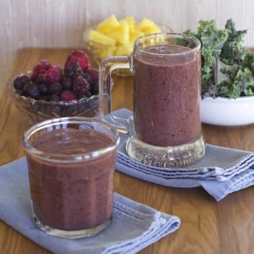 Two Triple Berry Kale Smoothies in mug and glass with fruit and kale in background