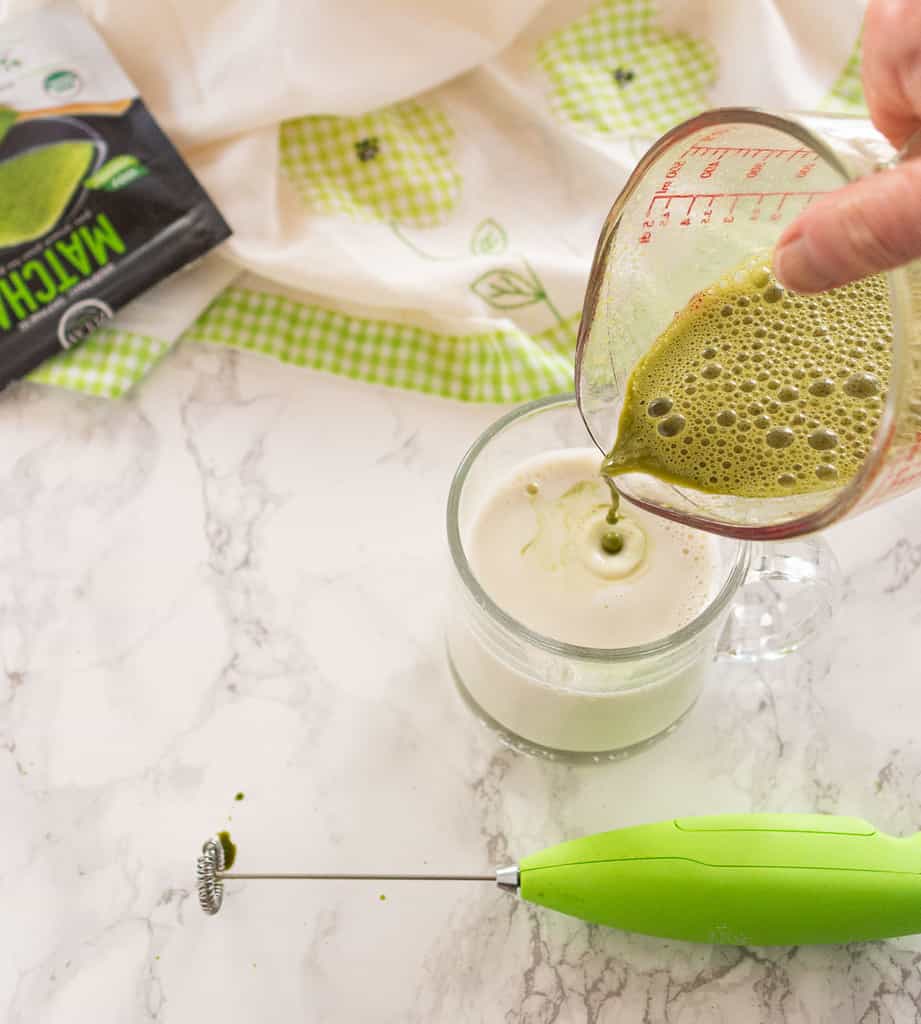 pouring matcha foam into mug of hot oat milk with green milk frother in foreground